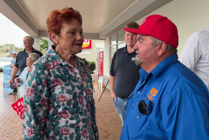 A woman with orange hair speaks with a man wearing a red hat and blue shirt under a building verandah