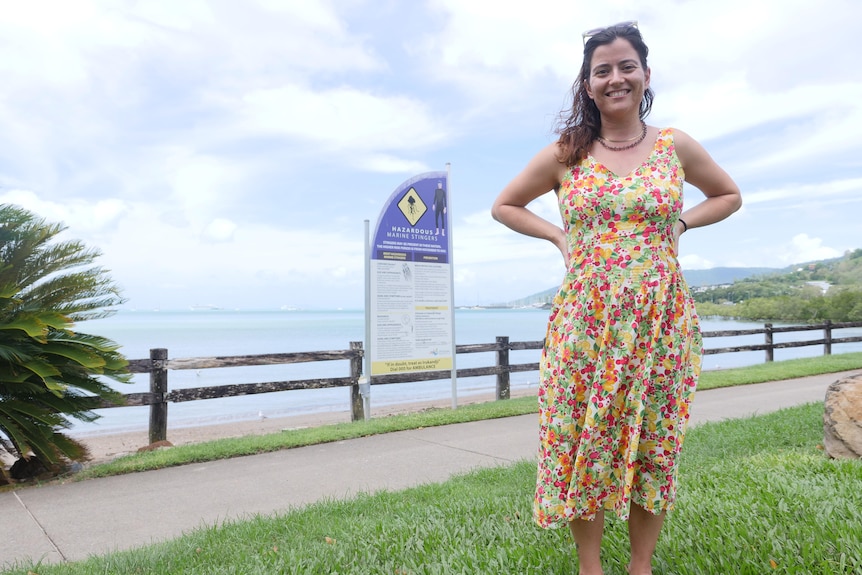 A woman in a floral dress stands smiling at the camera, with her hands on her waist. 