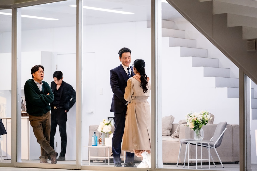 On stage, in a house with wall-to-ceiling windows, Doyeon Jeon, a Korean woman, speaks to a smiling Haesoo Park.