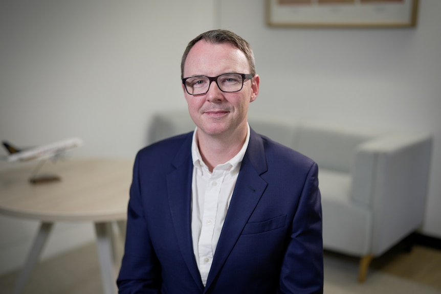 A man in a navy suit with a white shirt and glasses sits in an office space.
