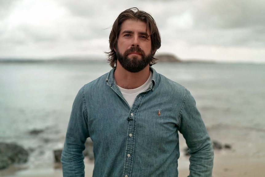 A man stands looking at the camera with a neutral expression. Behind him is a beach with water stretching the horizon.