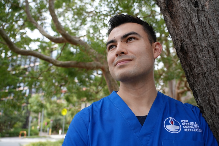 A man with short brown hair wearing blue scrubs standing under a tree.