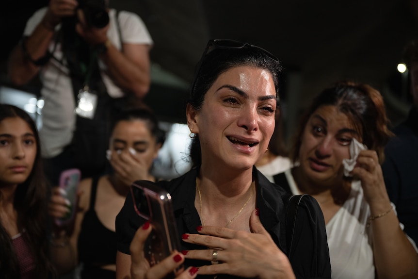 A woman holds a phone and cries, with her hand to her chest