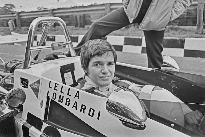 A black-and-white photo of a woman with short hair sitting in an old open racing car with "LELLA LOMBARDI" on the side.