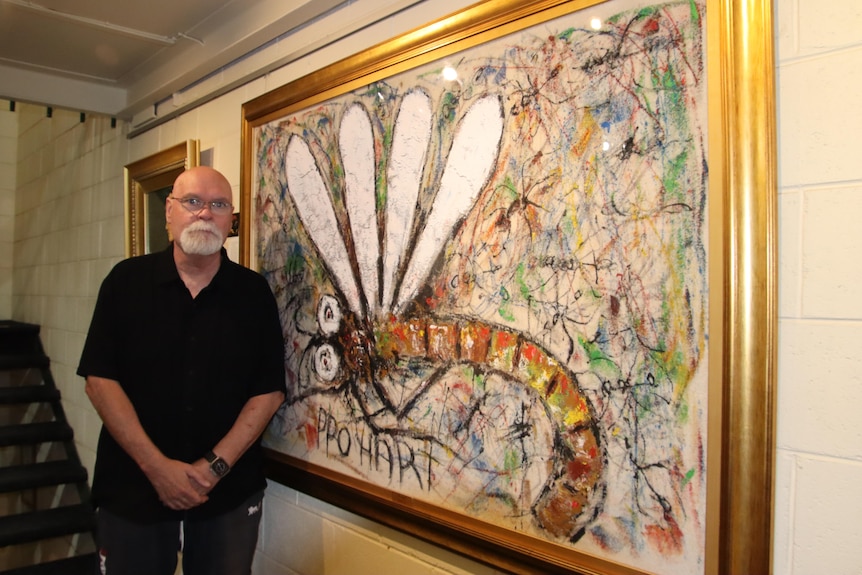 An older man in a black shirt standing next to a framed cut out piece of carpet with a colourful painting of a dragonfly on it.