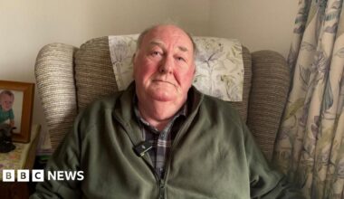 A man with grey hair sits in an arm chair wearing a checked shirt and a green zip up fleece. The arm chair is striped beige, with a patterned head rest that matches the curtain on the right of the picture. To the left, there is a picture of a young child on a side table.