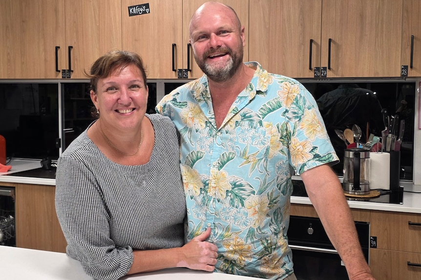 A couple - man and wife - stand together in a kitchen with their arms around eachother.