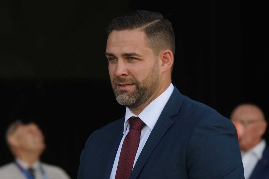 A man in a dark blue suit and a maroon tie looks into the distance.