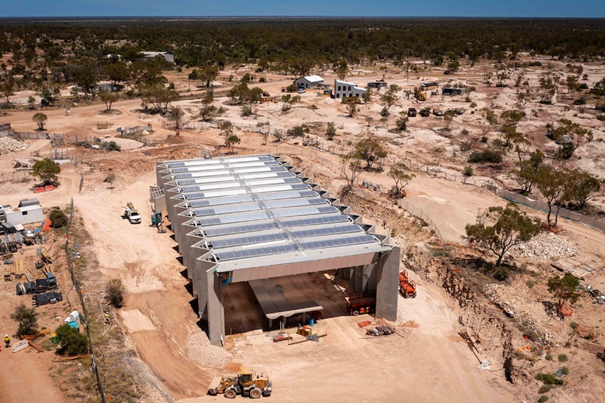 A half-built concrete structure in the shape of a rectangle is surrounded by a vast open plain of dirt and scrubby trees  