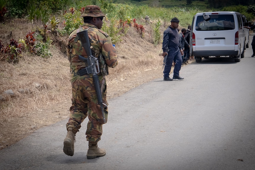 A man wearing army fatigues holds a gun while walking along a street.