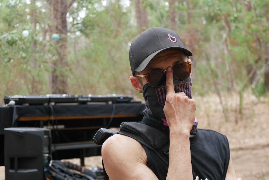 A man with a mask on and sunglasses standing in front of DJ decks.