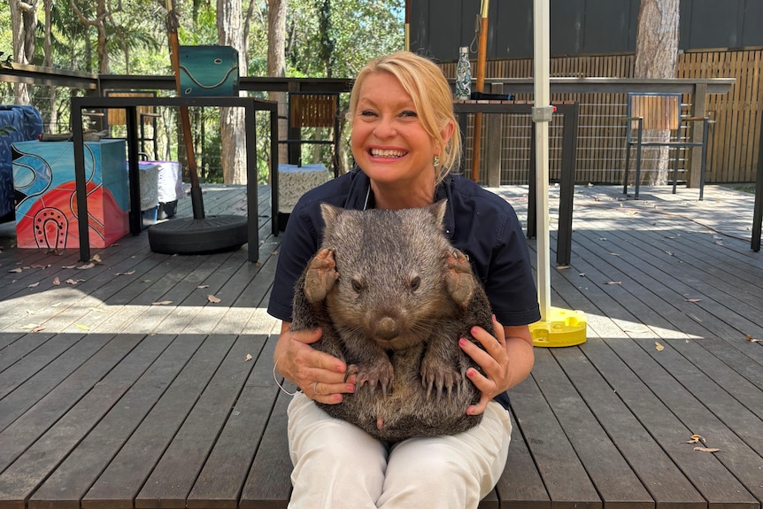 A blonde woman smiles at the camera with an adult wombat in her lap.