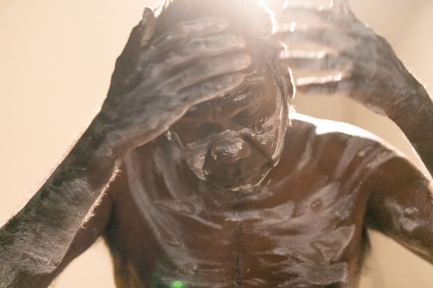 An Aboriginal man holds his hands up to his head as he paints himself white. Light shines behind.