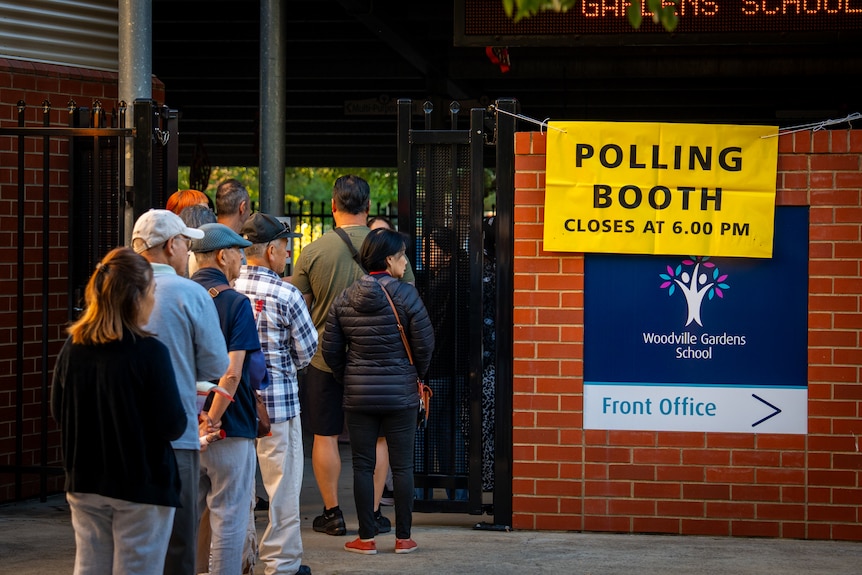 A line of voters at a gate near a wall that has a yellow sign reading 'Polling Booth closes at 6pm'