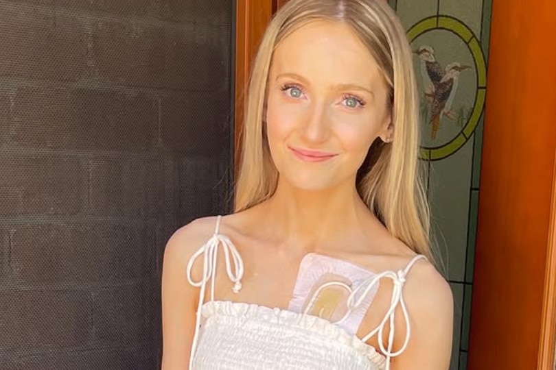 A young woman with blonde hair and a medical tube visible underneath her white top.