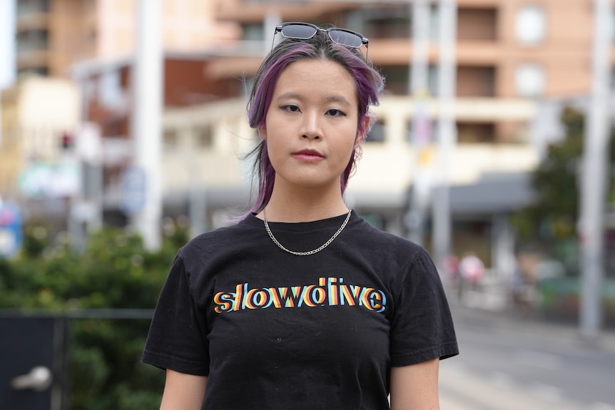 Ms Wang with purple-coloured hair, a necklace and sunnies on her head stands outside looking at the camera.