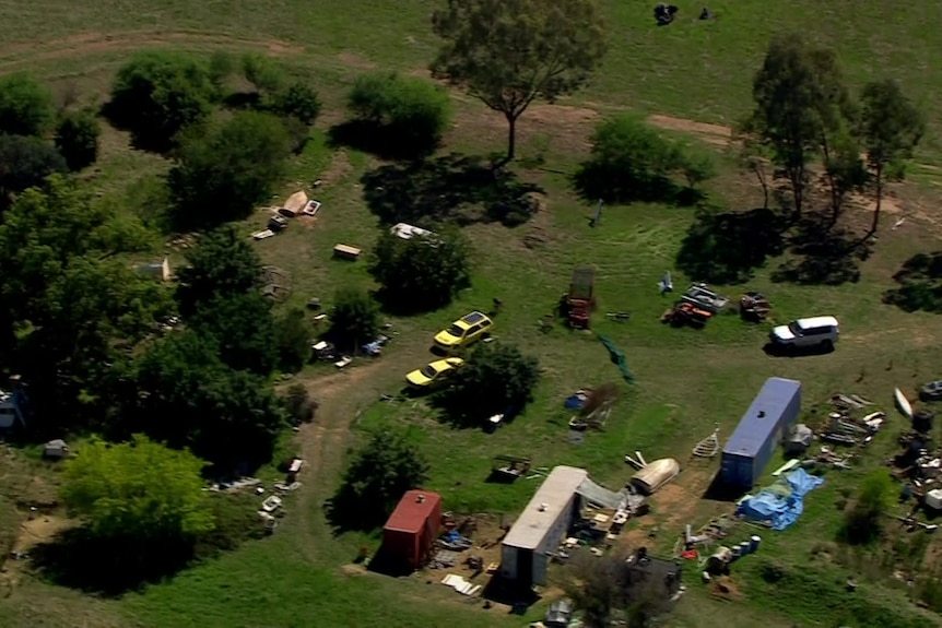 Two shipping containers, old cars and scrap metal are scattered amongst trees on a rural property.