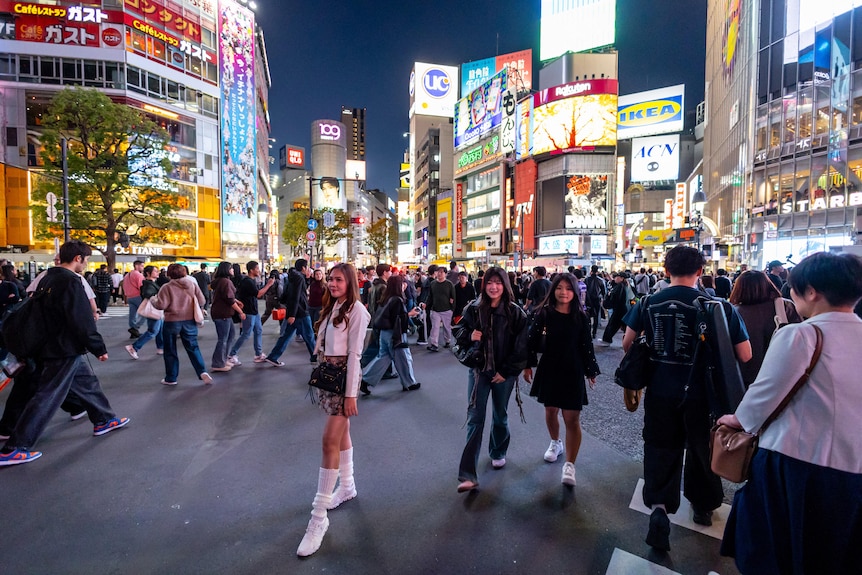 A young woman poses for a photo as a large crowd of people cross a bright, colourful neon lit intersection.