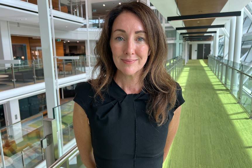 Mary stands in an office corridor, wearing a sleeveless black dress, with long hair.