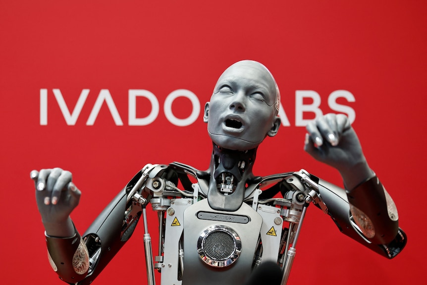 A robot with a human-like face with its arms held high. Behind it is a red backdrop with white writing.