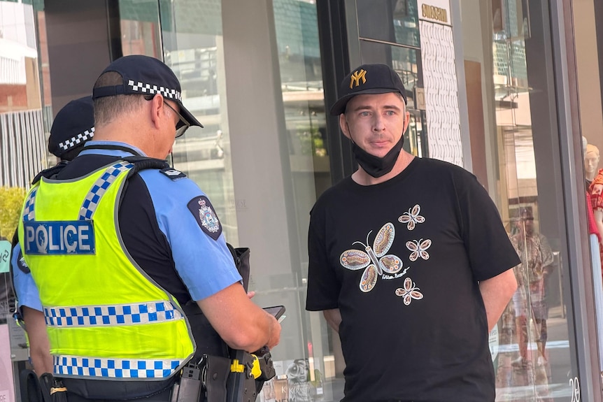 Police speak to a man wearing a black butterfly print T-shirt, baseball cap and face mask