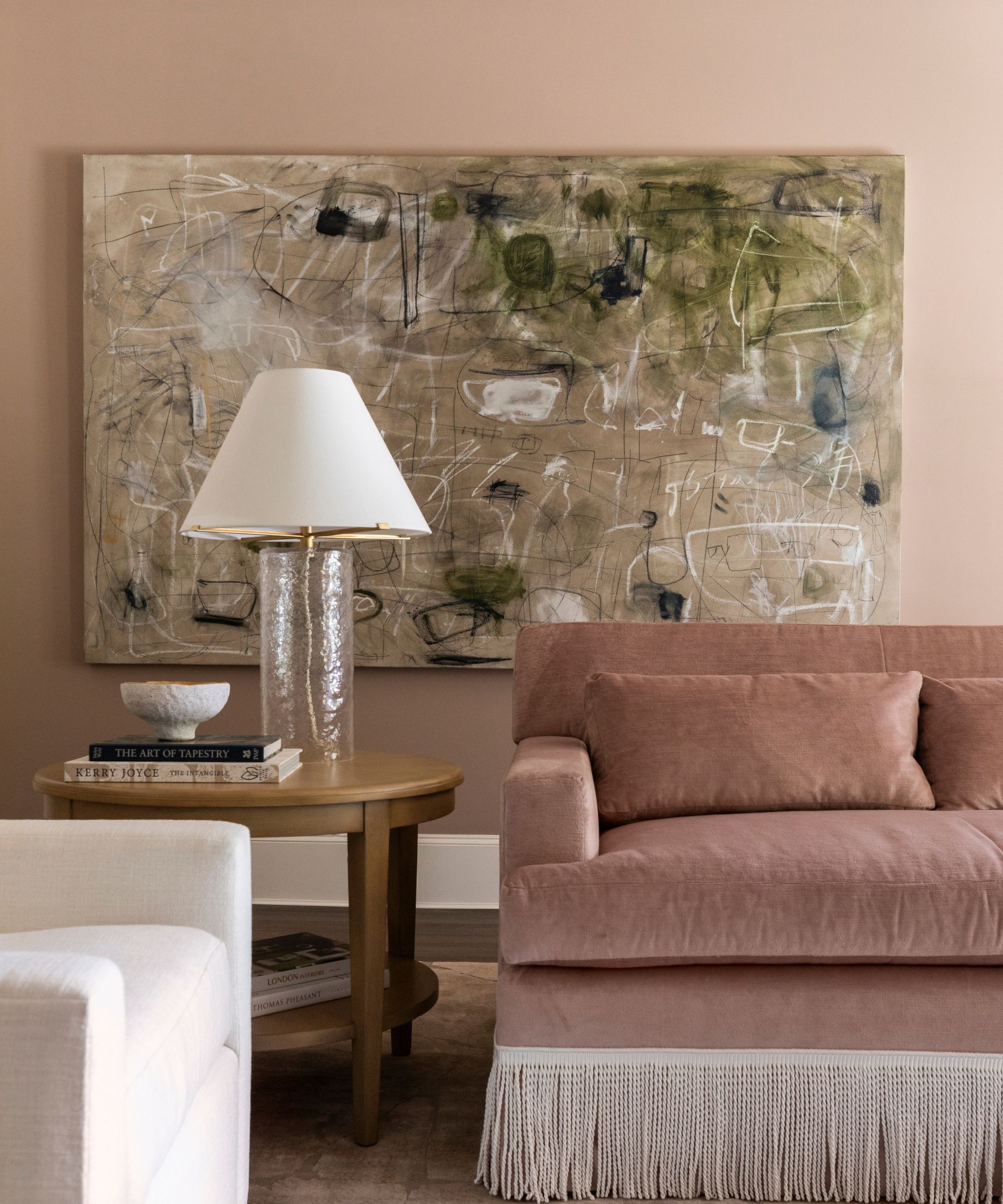 A traditional living room with a dusky pink sofa with a fringe edge, muted pink walls, a wooden side table with a cream lamp, and a large abstract piece of artwork on the wall.