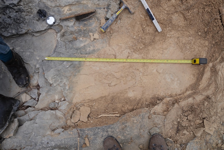View of rock and dirt ground with tape measure stretched out and brush tools, two sets of shoes looking on