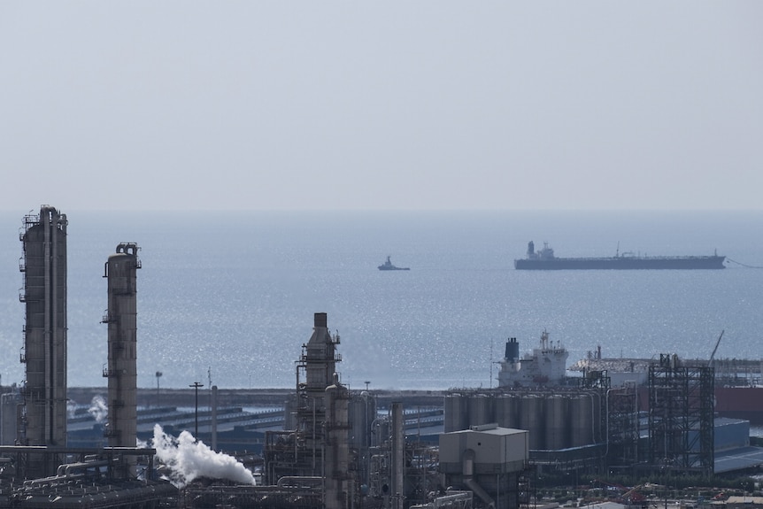 A wide view of gas infrastructure by the sea. A large ship is sailing through the ocean.