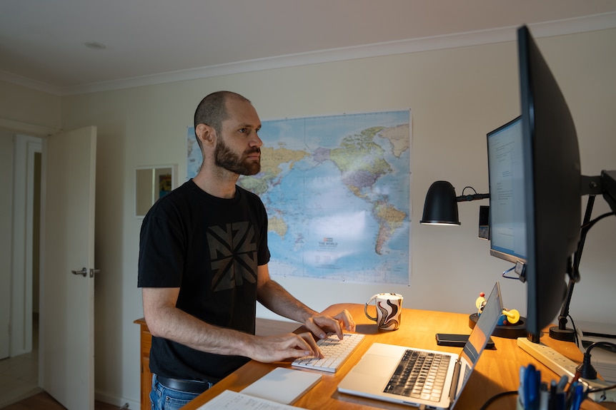 A serious looking man with closely shaved head and beard stands working in a home office set-up.