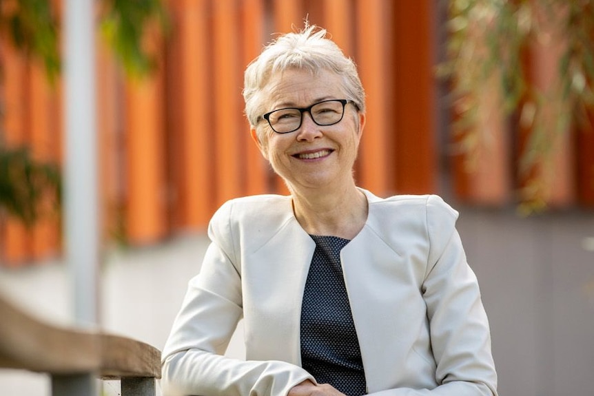 A woman with silver-white hair and glasses looks at the camera and smiles