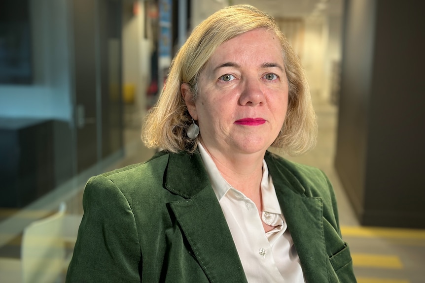 Profile photo of a woman with short blond hair in a green blazer and white shirt and red lipstick.