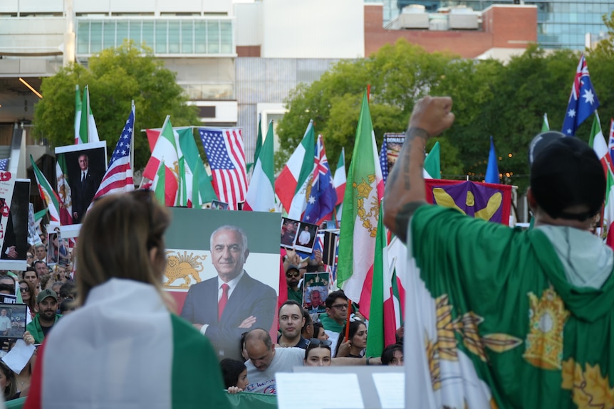 Crowds of people are holding flags and posters, as they look up at a stage. Two speakers have flags around their shoulders.