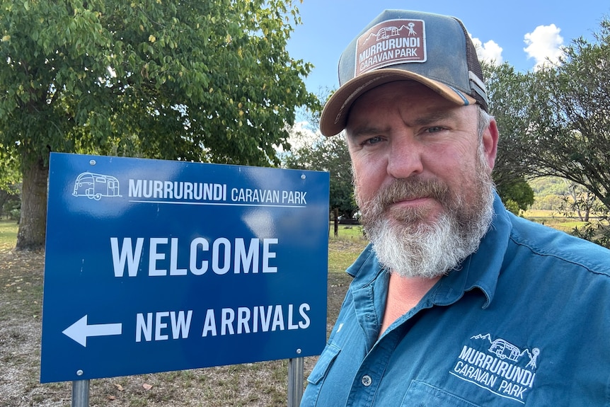 A man in a cap stands in front of a sign saying Welcome New Arrivals.