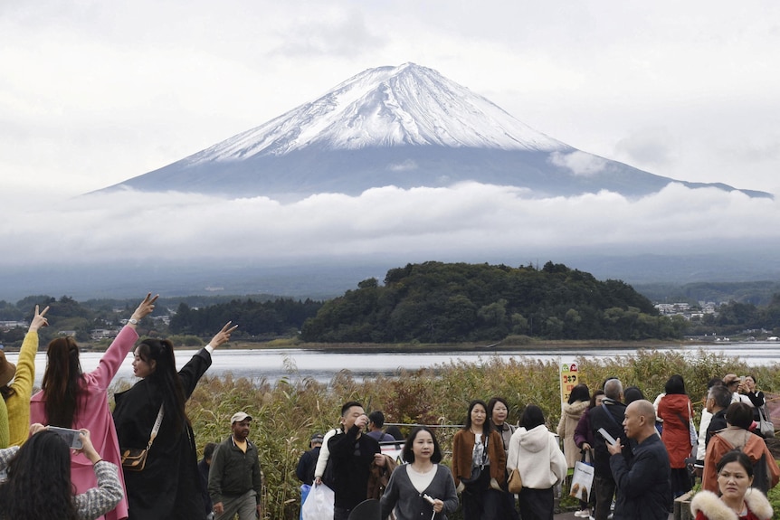 Tourists take photos and pose at a lookout with a clear and close view of Mount Fuji covered in snow.