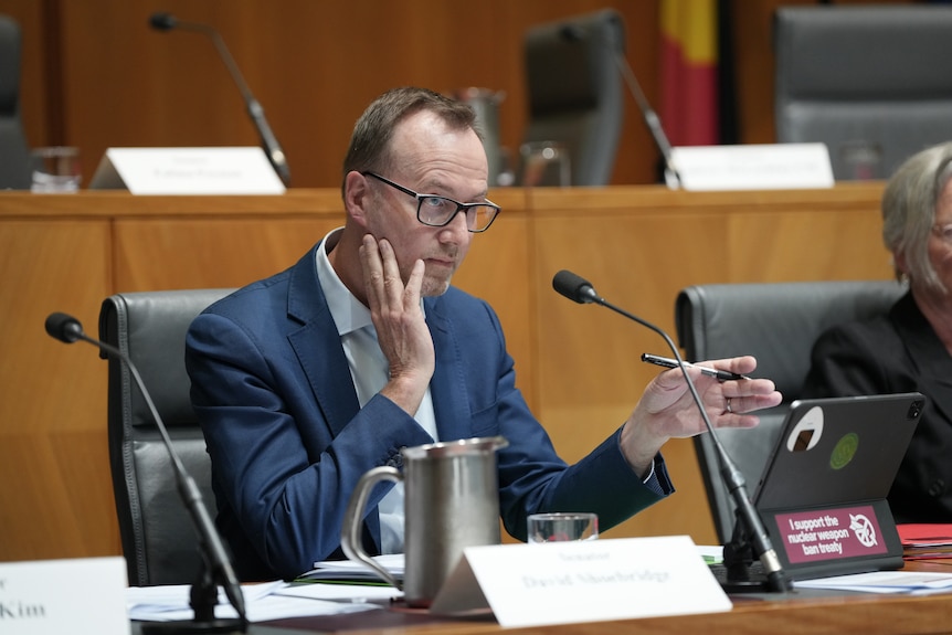 A man at a desk, wearing a navy suit and glasses. 