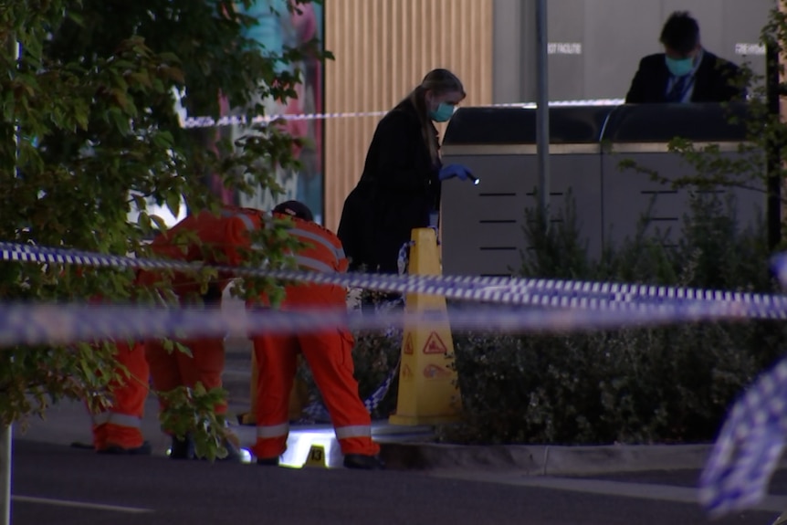 Two people in orange overalls bend over something on the ground as two people in dark clothing look in a bin behind police tape.