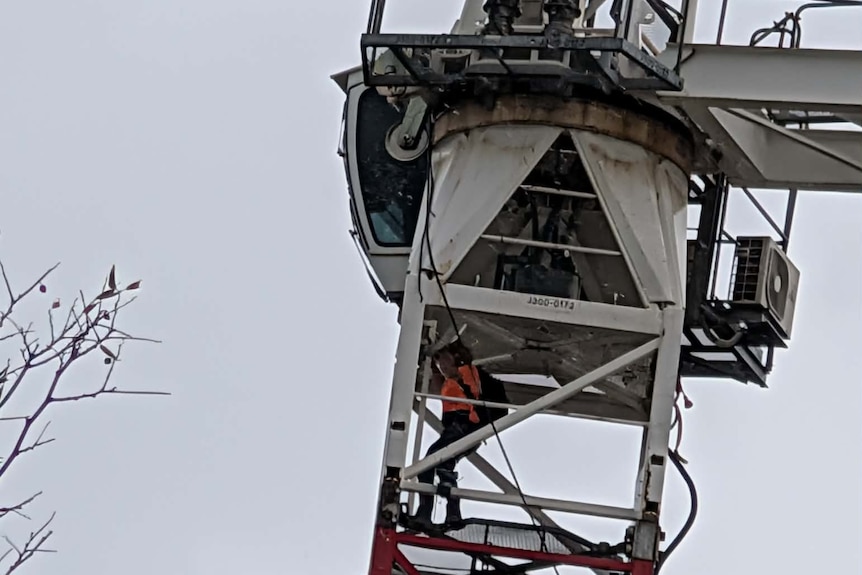 A woman wearing a hi-vis orange shirt wearing a backpack descends down a ladder underneath the crane cabin.