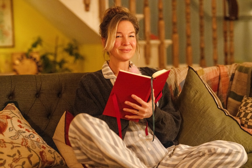 Renée Zellweger smiles in the latest Bridget Jones film, perched on a couch in pajamas  with her trademark red diary.