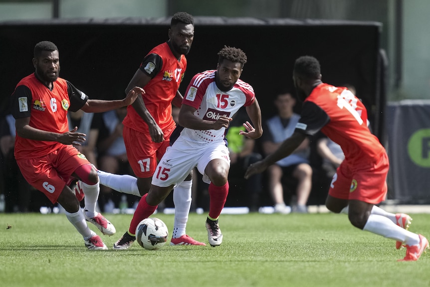 A man in white & red footy uniforms makes aplay with a soccer ball amidst 3 other soccer players wearing all red soccer uniforms