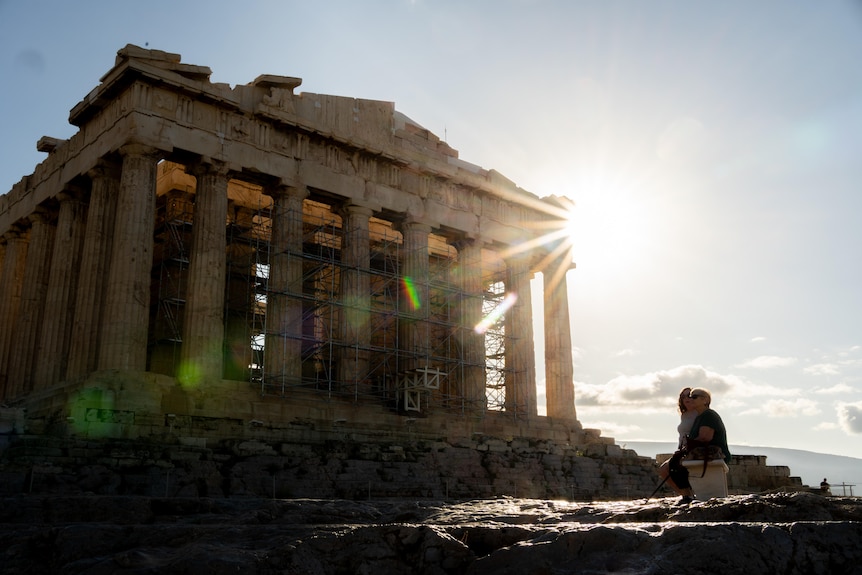 Two women seated in front of the Parthenon, which is surrounded by scaffolds.