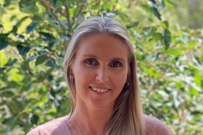 A woman with long blonde hair smiles in front of green foliage.