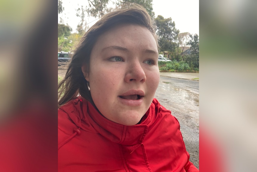 A close-up shot of a teenage girl with brown hair, wearing a red rain jacket in front of a road and trees.