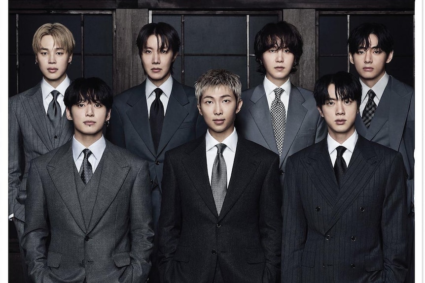 Seven members of K-pop group BTS pose in dark-grey business suits in a line