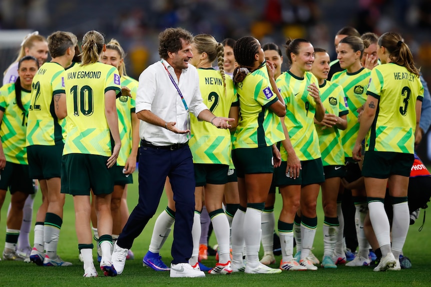 An Australian coach walks around on the pitch after a match, while Matildas players relax after the game.