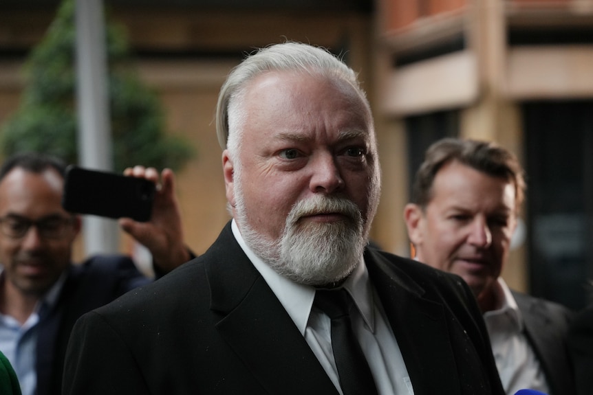 Kyle Sandilands has white hair and a beard and is surrounded by reporters.