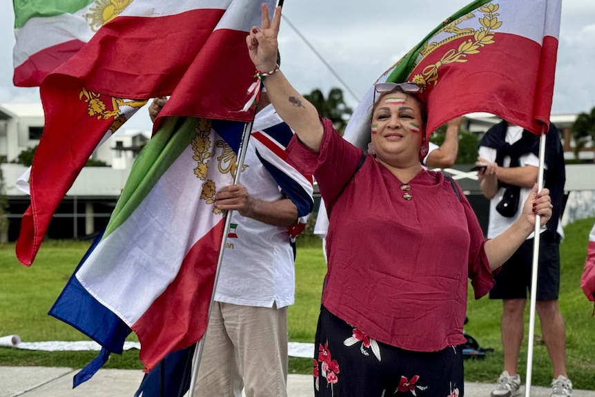 A woman with a red shirt holds an Iranian lion and sun flag and flashes a peace sign
