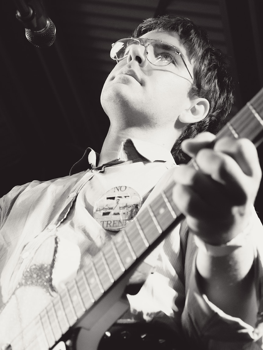 Black and white photo of man wearing glasses and shirt holding guitar on stage
