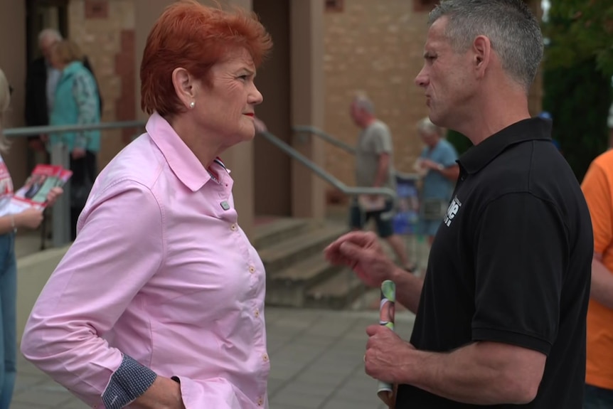 A woman with orange hair speaks to a man wearing a black shirt