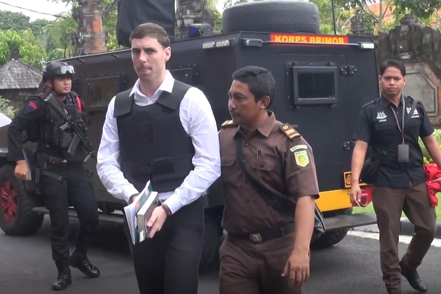 A man wearing a white shirt and black vest grimaces as he is escorted by a police officer.