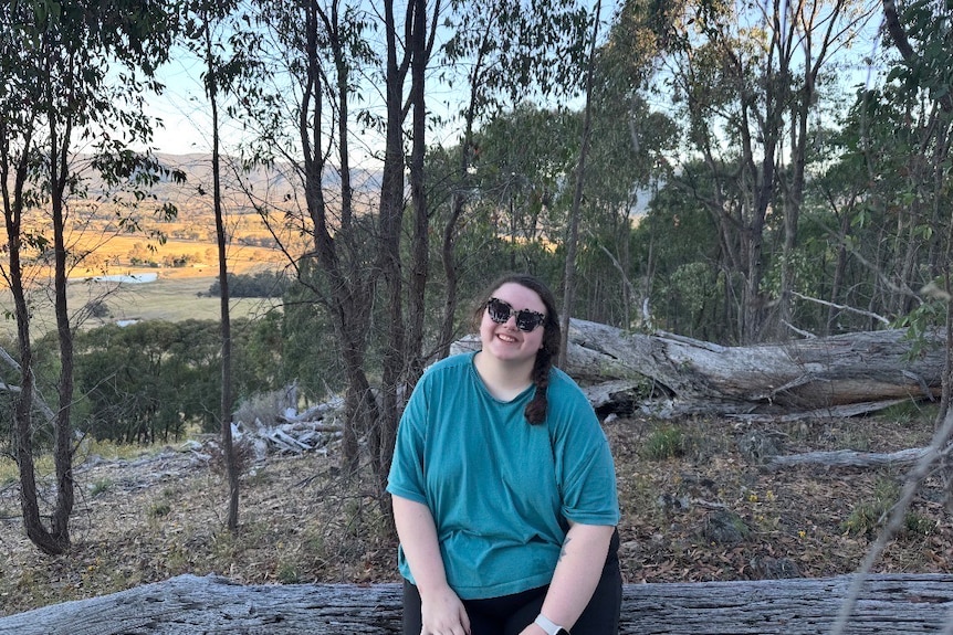 Rachel sits on a tree log in country Victoria smiling at the camera.